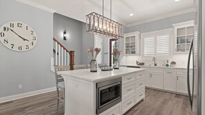A modern kitchen with white cabinetry, a central island with a built-in oven, a large wall clock, and a unique pendant light. - New Home in King City, Sleeps 10, Pool and Screened in Porch with TV! (St Simons Island)