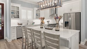 A modern kitchen with white cabinetry, a stainless steel refrigerator, and a central island with bar stools. - New Home in King City, Sleeps 10, Pool and Screened in Porch with TV! (St Simons Island)