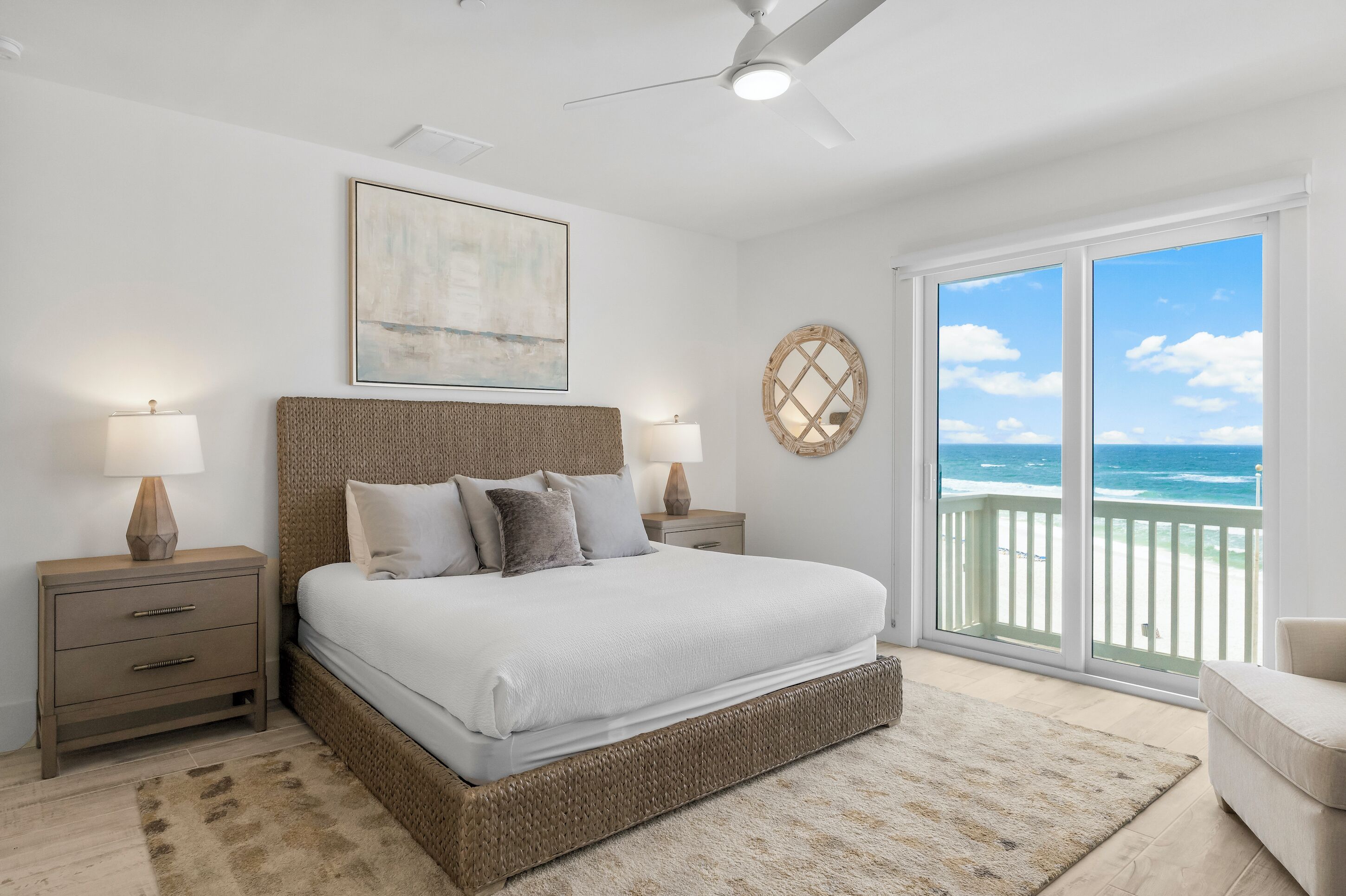 A bedroom with a large bed, two bedside tables with lamps, a ceiling fan, a large window with a view of the ocean, and a framed artwork on the wall.
