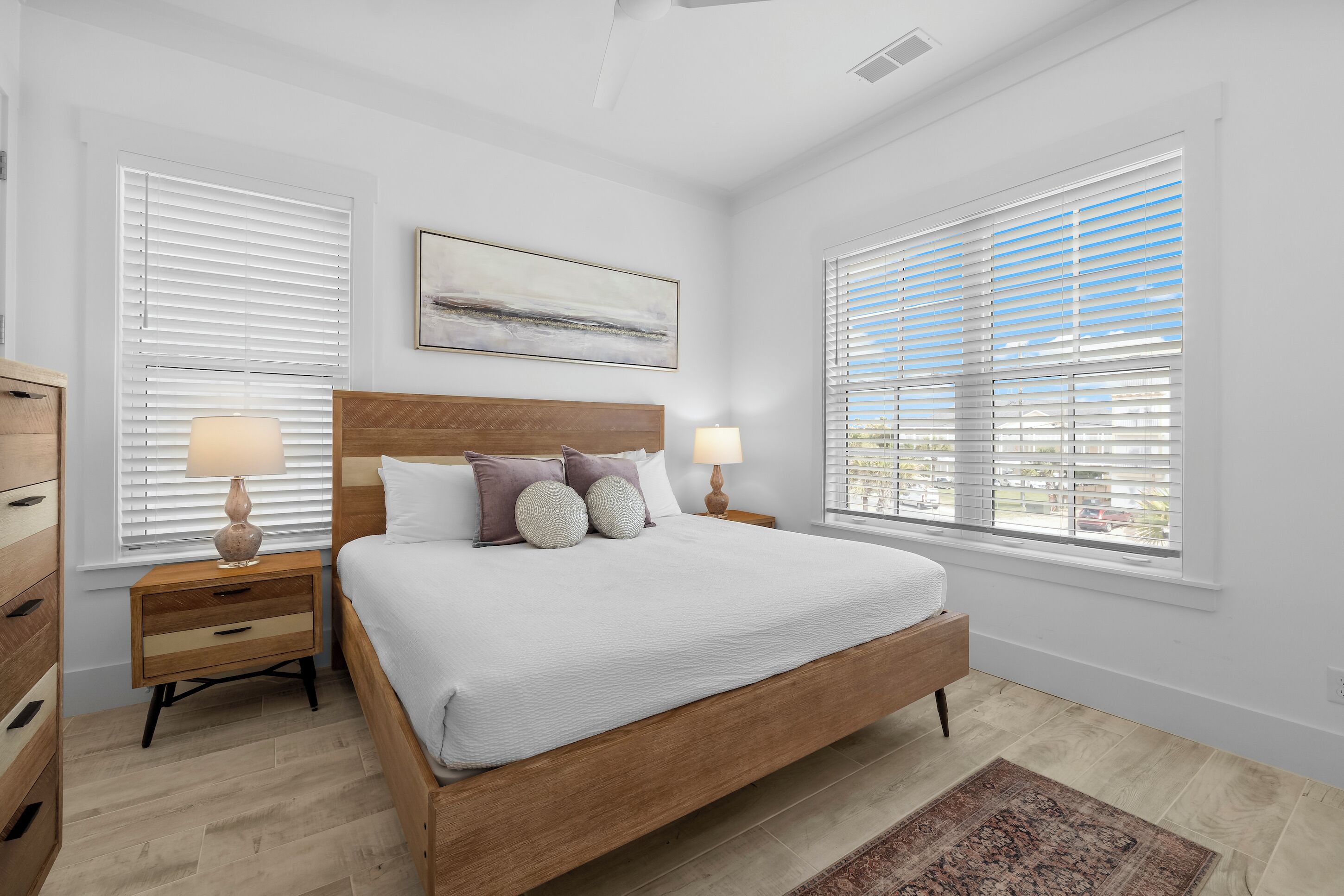 A bedroom with a wooden bed, two bedside tables with lamps, a large window with blinds, and a painting on the wall.