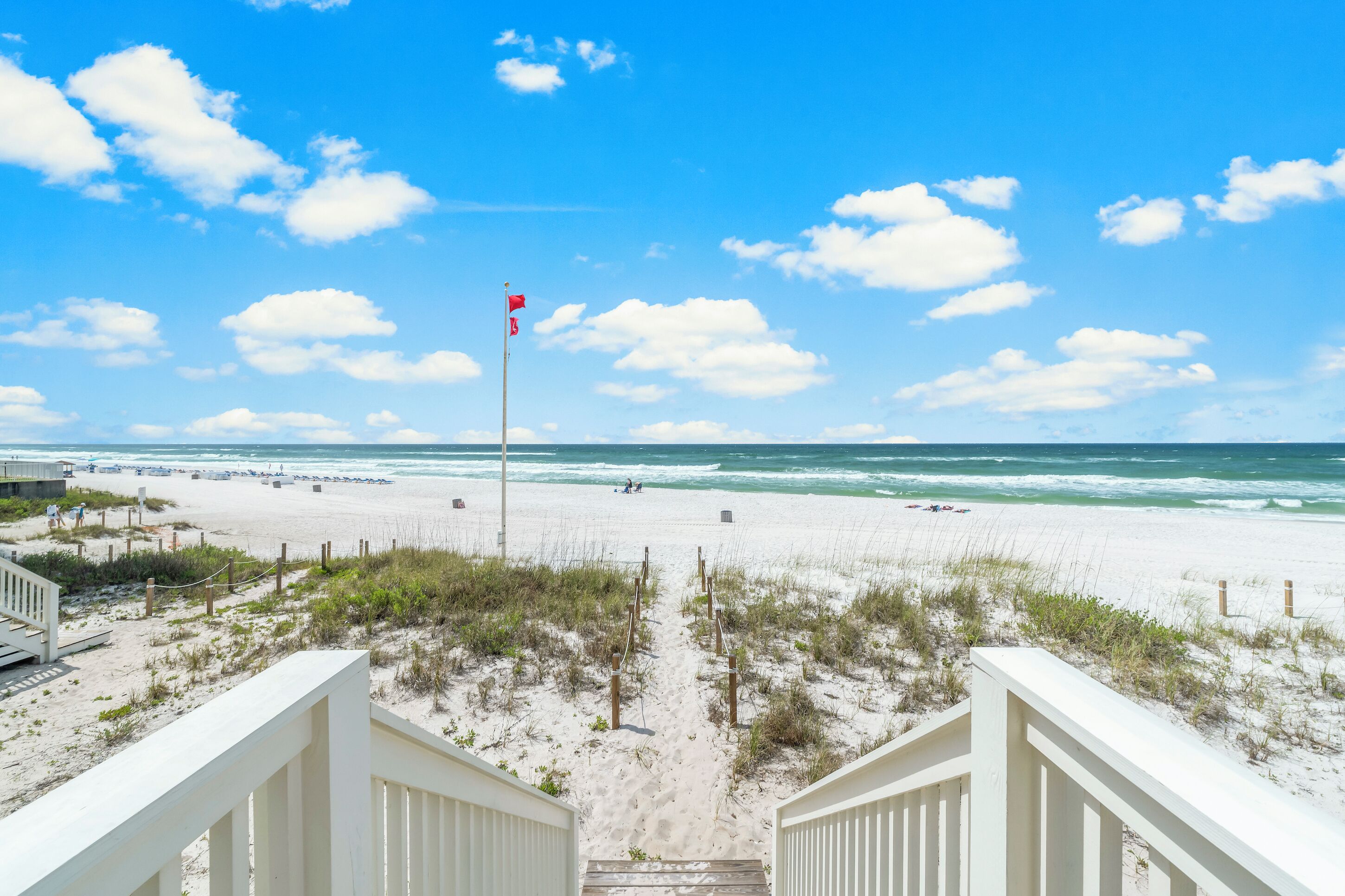 Une vue sur la plage avec un escalier en bois descendant jusqu'au sable, un mât avec un drapeau, et l'océan en arrière-plan.