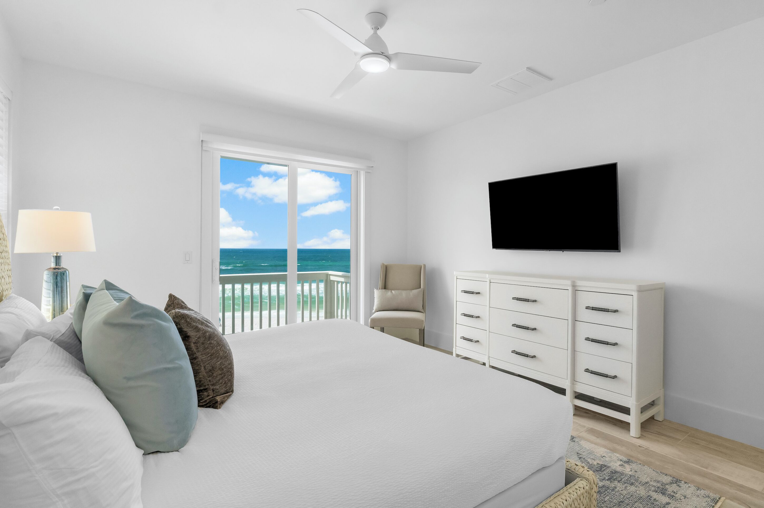 A bedroom with a bed, a TV, a chair, a dresser, and a balcony with a view of the ocean.