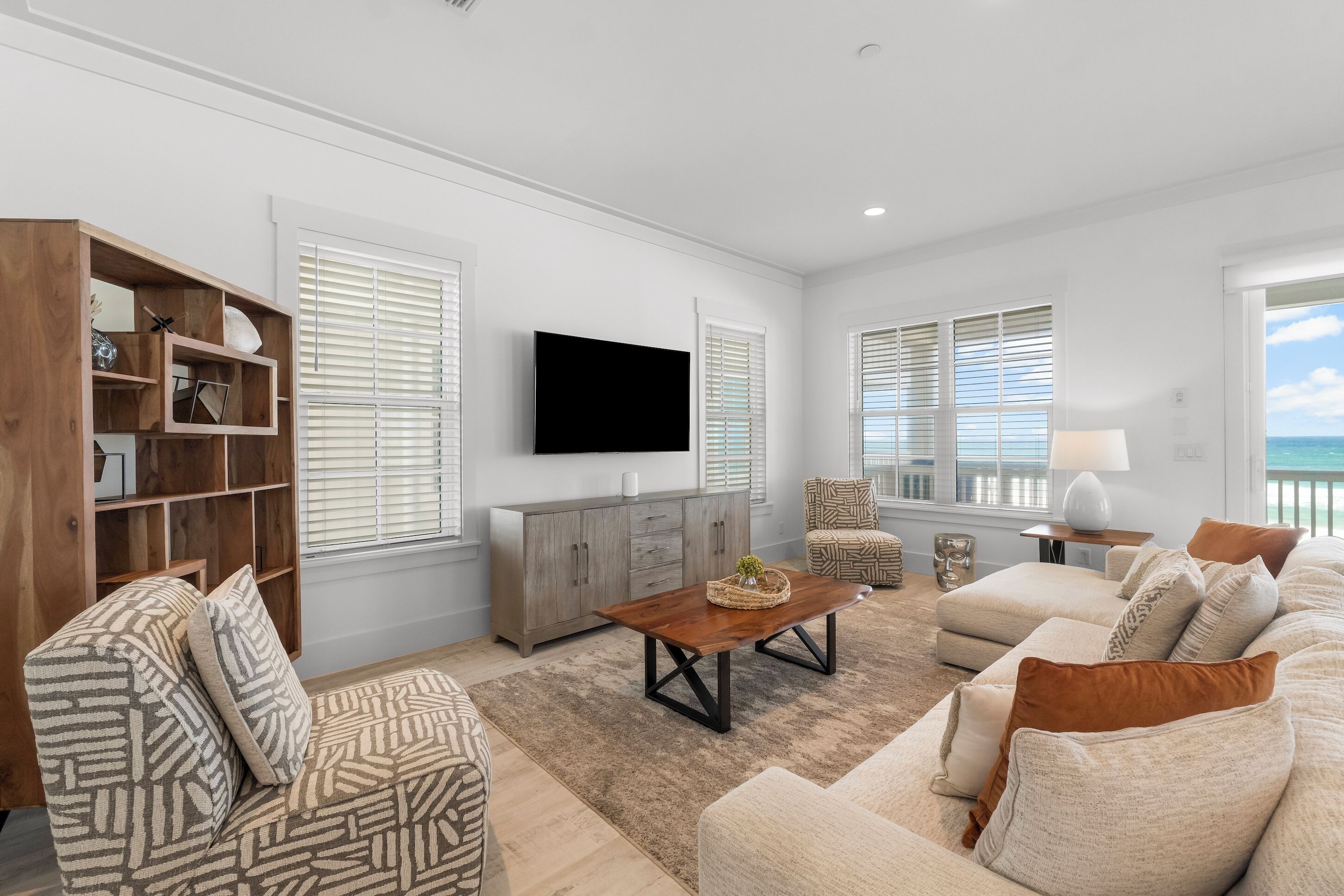 A modern living room with a large flat-screen TV, a wooden entertainment unit, a beige sofa set, and a view of the ocean through large windows.