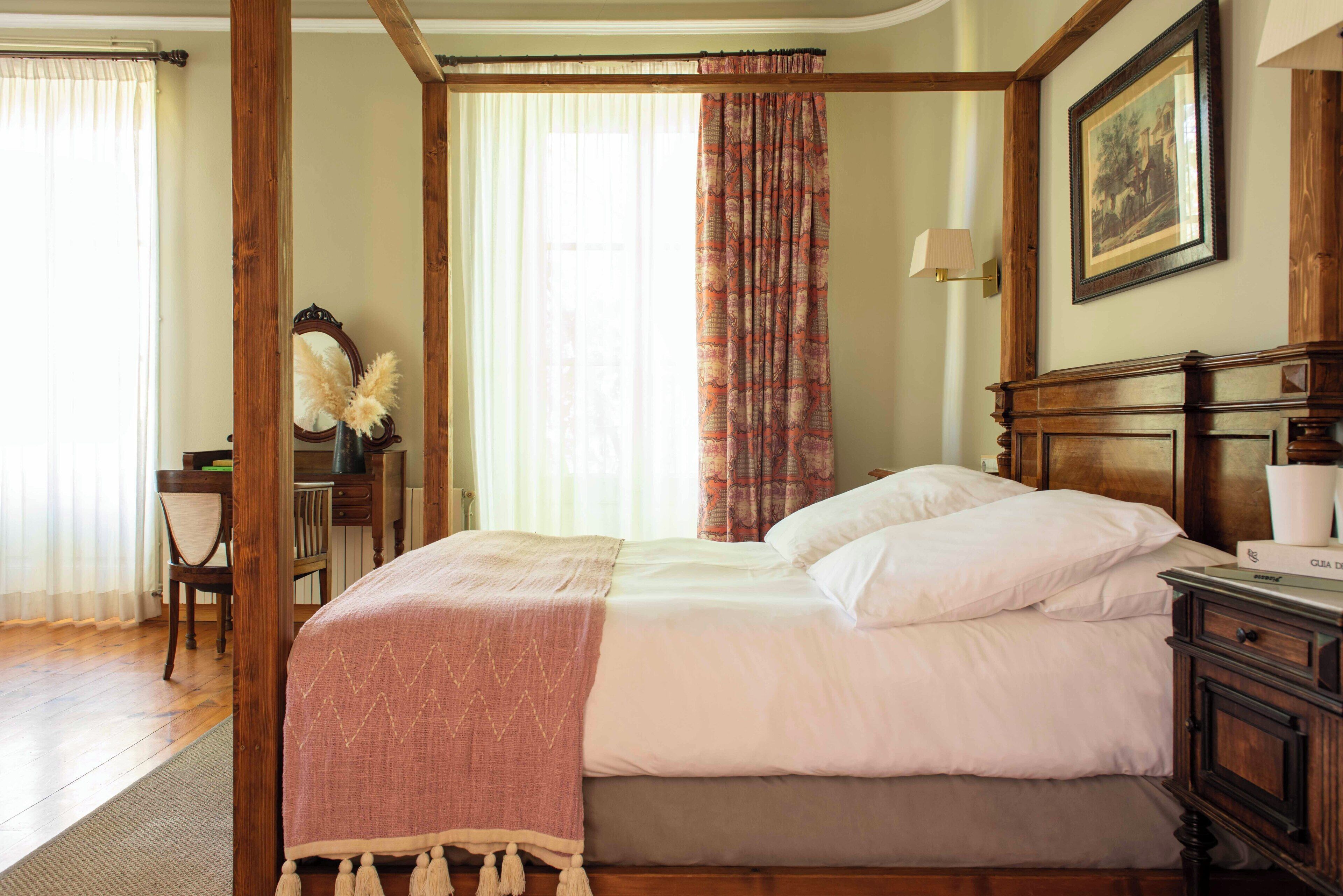 Een slaapkamer met een hemelbed van hout, een bureau met stoel, een spiegel en een ingelijste foto aan de muur.
