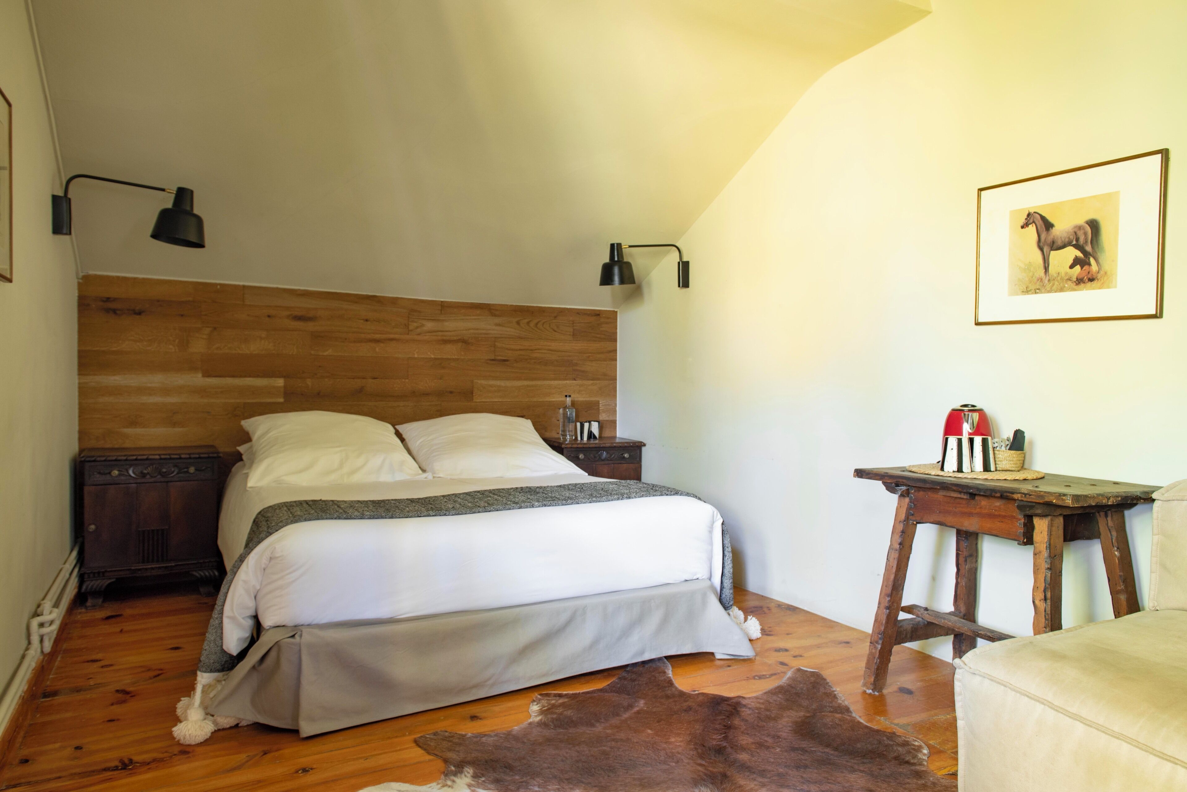 A bedroom with a wooden headboard, a bed with white linens, a wooden nightstand, a small table with a lamp, and a framed picture on the wall.
