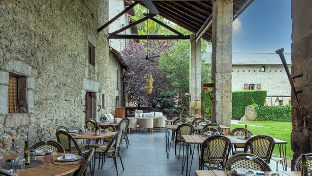 An outdoor patio with stone walls, wooden tables, wicker chairs, and a view of a garden and building in the background. - Mas Sant Marc (Puigcerda)