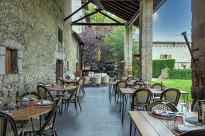 An outdoor patio with stone walls, wooden tables, wicker chairs, and a view of a garden and building in the background. - Mas Sant Marc (Puigcerda)