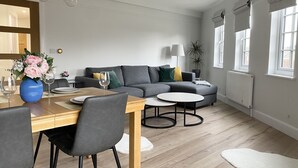 Una sala de estar moderna con un sofá gris, una mesa de comedor con sillas y un amplio ventanal con cortinas.