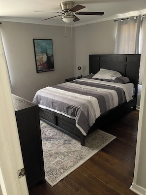 Un dormitorio con cama, ventilador de techo, mesita de noche, lámpara y un cuadro en la pared.