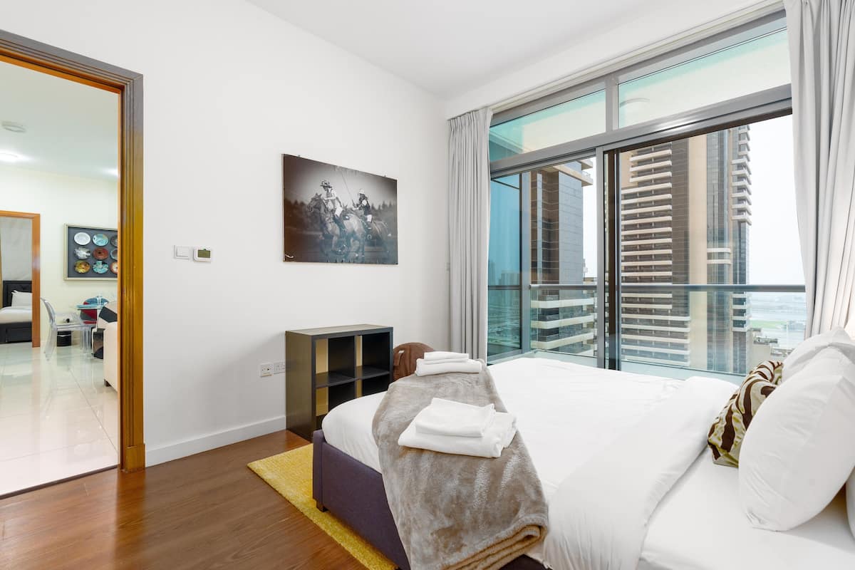 A modern hotel room with a large bed, a glass wall with a city view, a painting on the wall, and a small seating area.