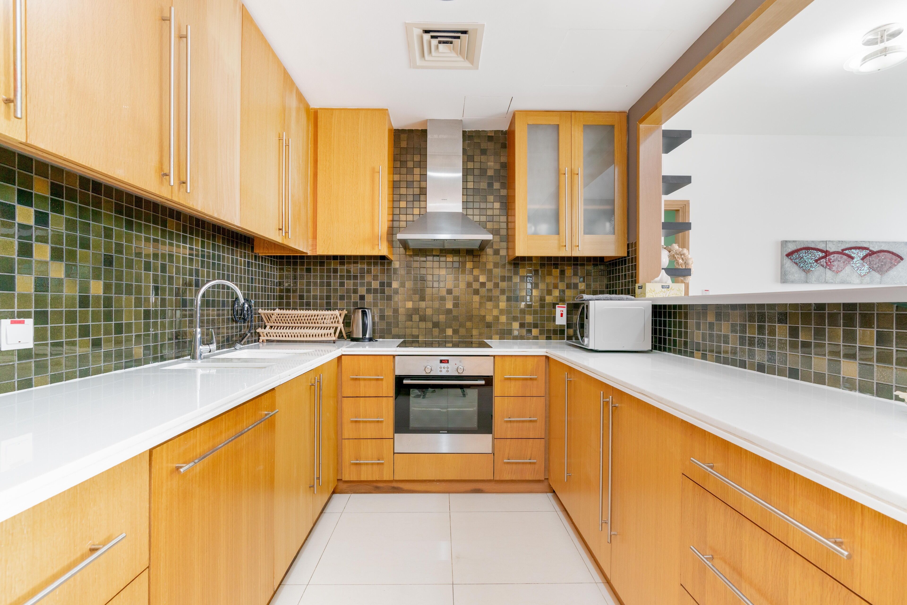 A modern kitchen with wooden cabinets, a stainless steel range hood, a microwave, and a tiled backsplash.