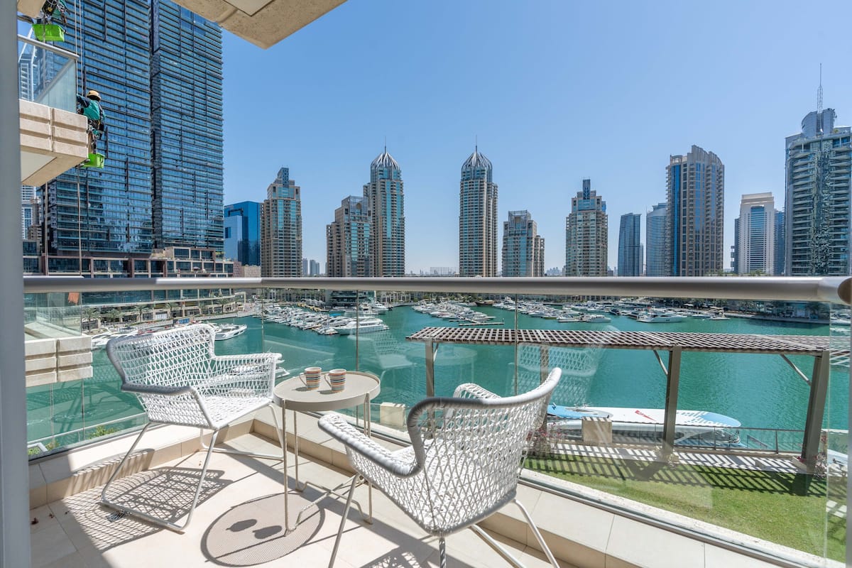 A balcony with white wicker chairs and a small table, overlooking a marina with boats and tall buildings in the background.