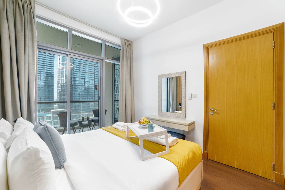 A hotel room with a bed, a bedside table with a bowl of fruit, a mirror, and a view of the city through large windows.