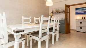 Un comedor con una mesa y sillas blancas, luces colgantes, una puerta de madera y un cuadro en la pared.