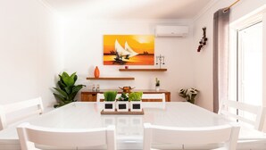 Een eethoek met een witte tafel en stoelen, een schilderij van een zeilboot aan de muur, en een potplant aan de linkerkant.