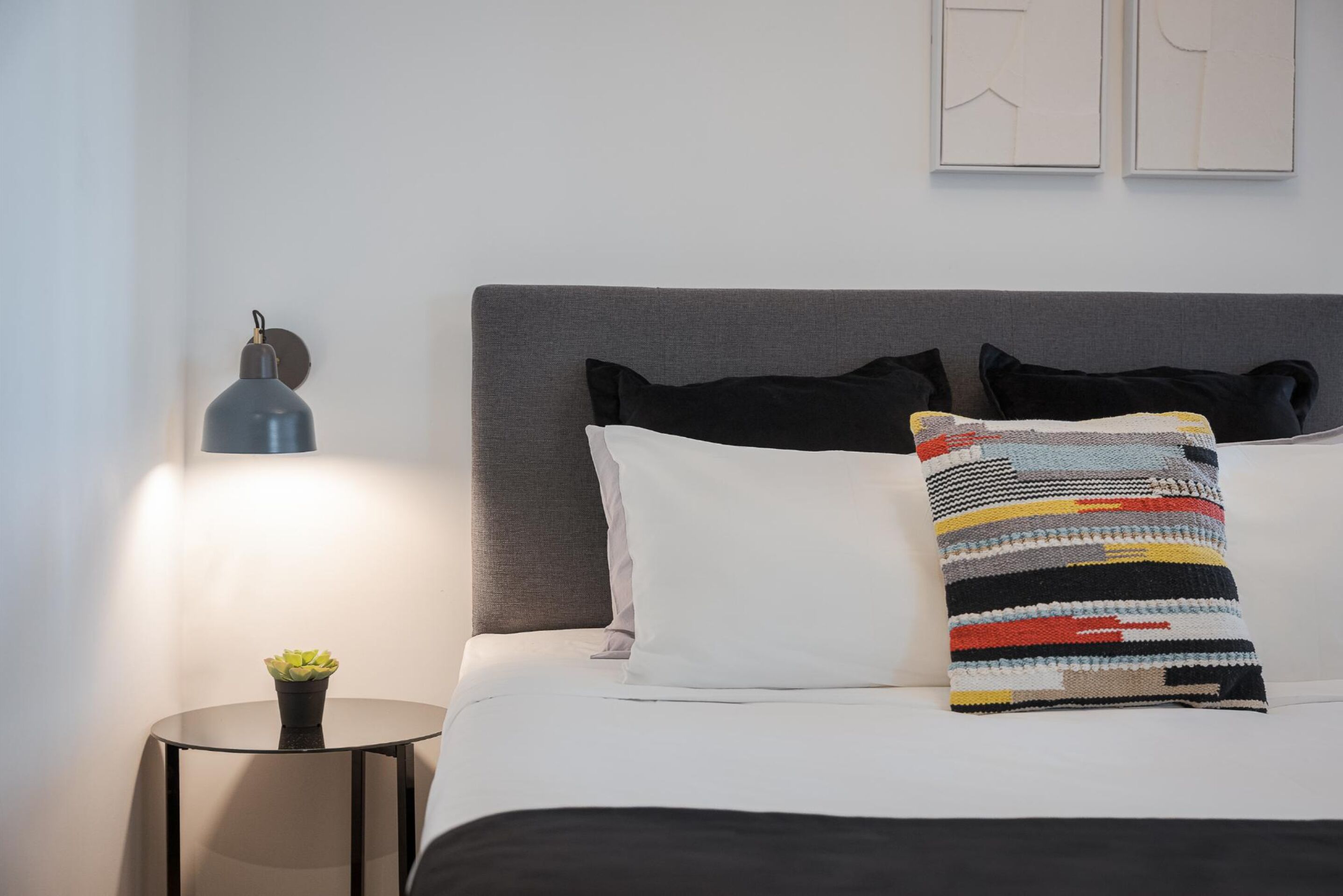 Ein ordentlich bezogenes Bett mit grauem Kopfteil, weißen und schwarzen Kissen sowie einem farbenfrohen Kissen mit Streifen. Neben dem Bett steht ein kleiner Tisch mit einer Lampe und einem Blumentopf.