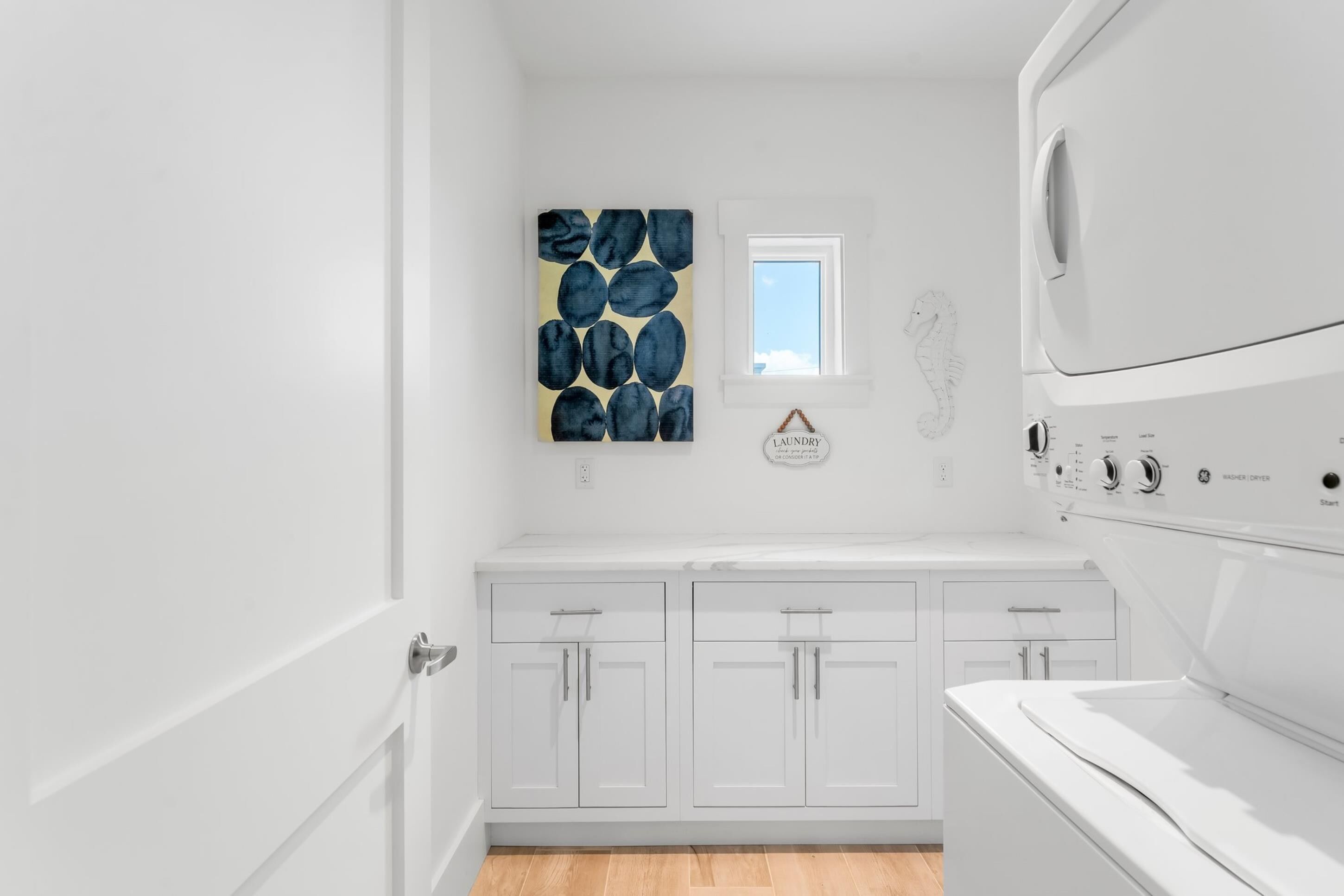 A laundry room with white cabinetry, a large wall-mounted washer and dryer, a seahorse decoration, and a blue abstract painting.