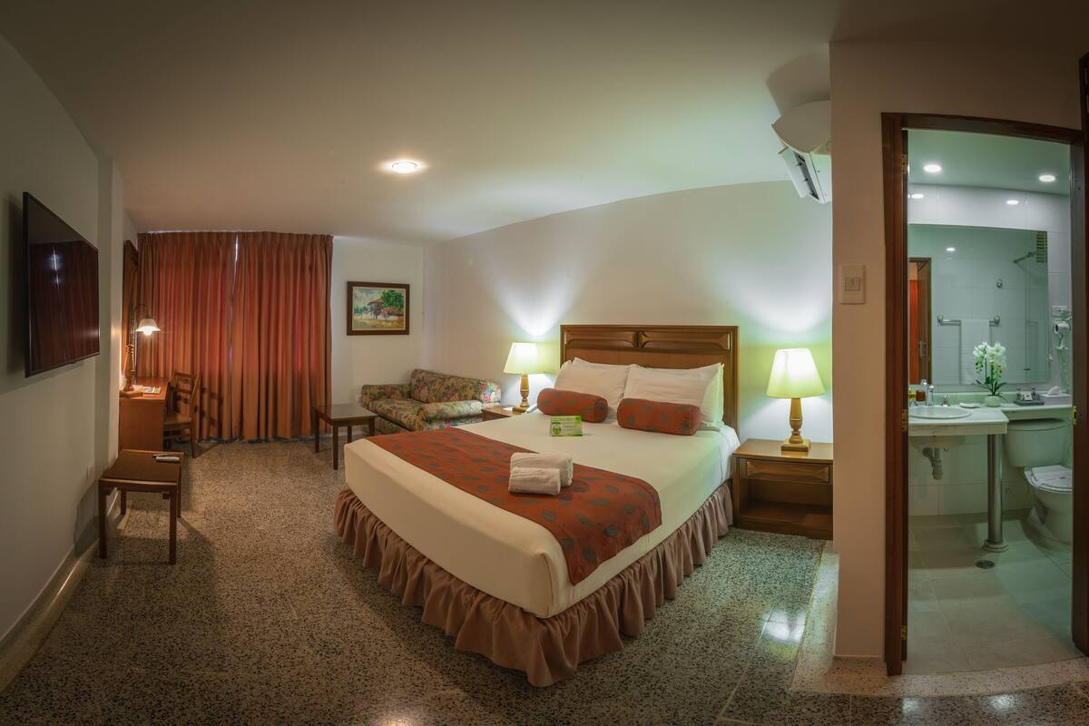 A hotel room with a bed, bedside table, lamp, chair, and a view of the bathroom.