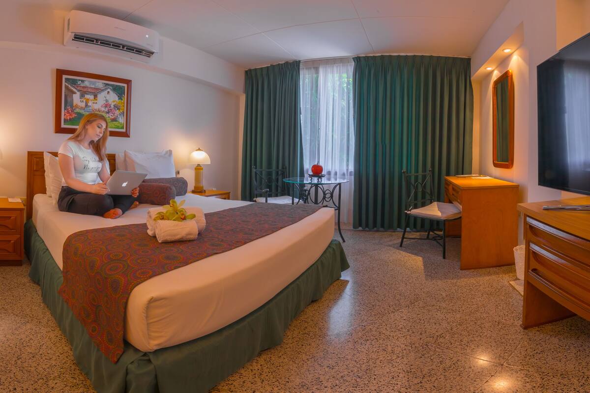 A hotel room with a bed, a desk, a chair, a TV, and a painting on the wall.