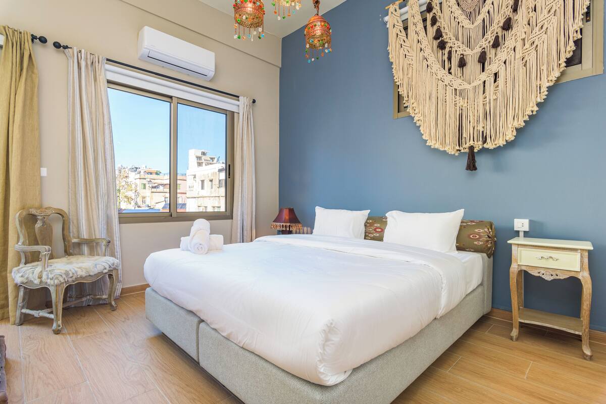 A bedroom with a large bed, a chair, a nightstand, and a macrame wall hanging.