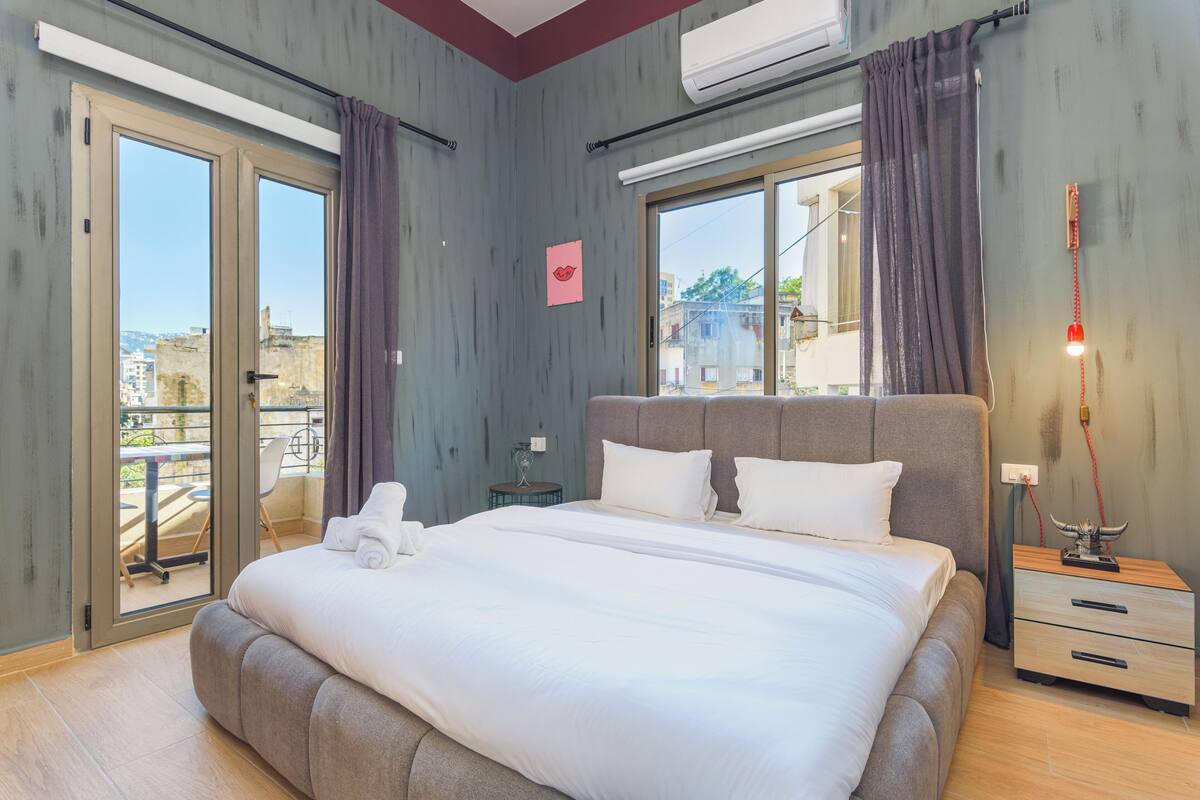 A hotel room with a large bed, a bedside table, a lamp, and a view of the city through the windows.
