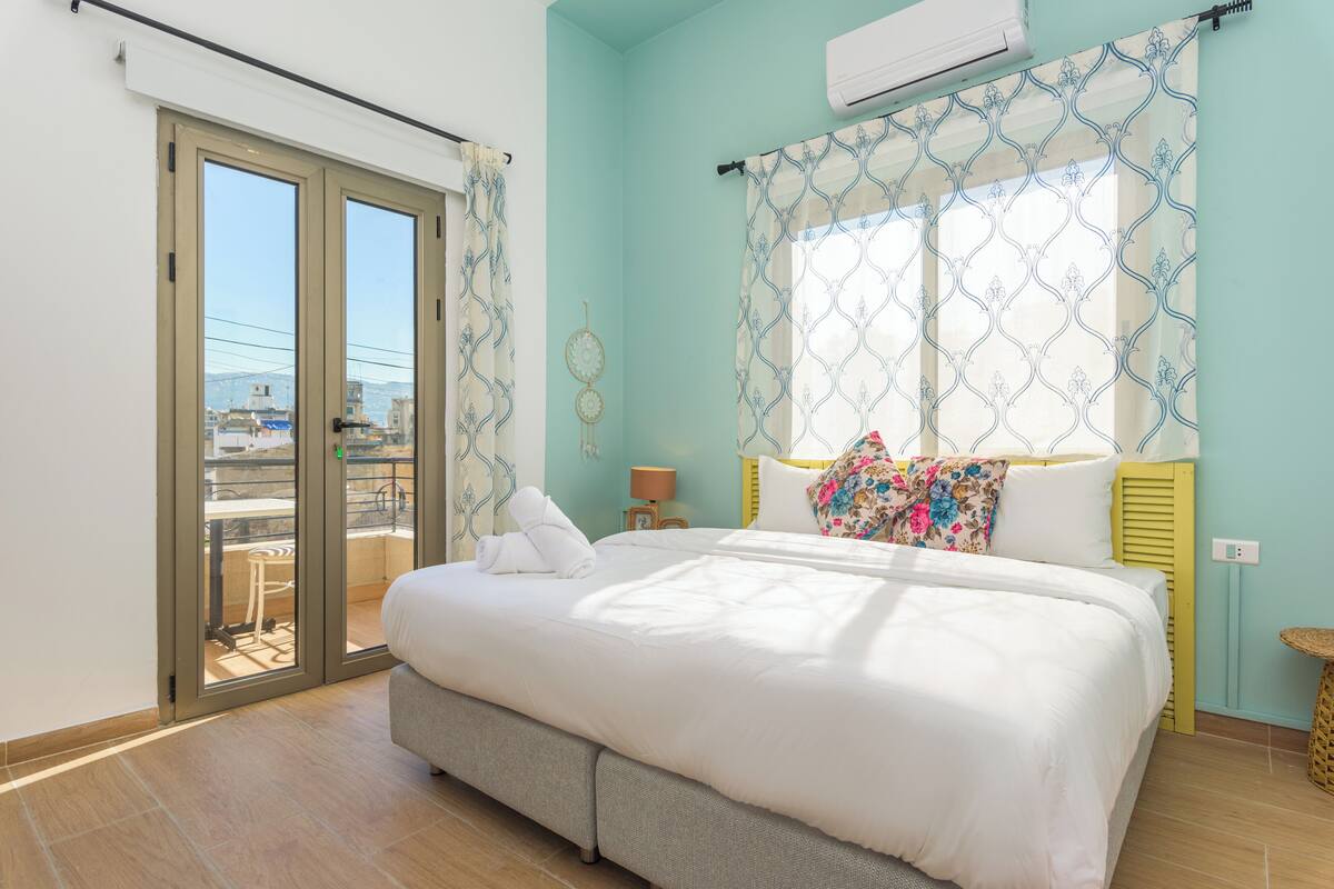 A bedroom with a bed, a window with patterned curtains, a bedside table with a lamp, and a view of the outside.