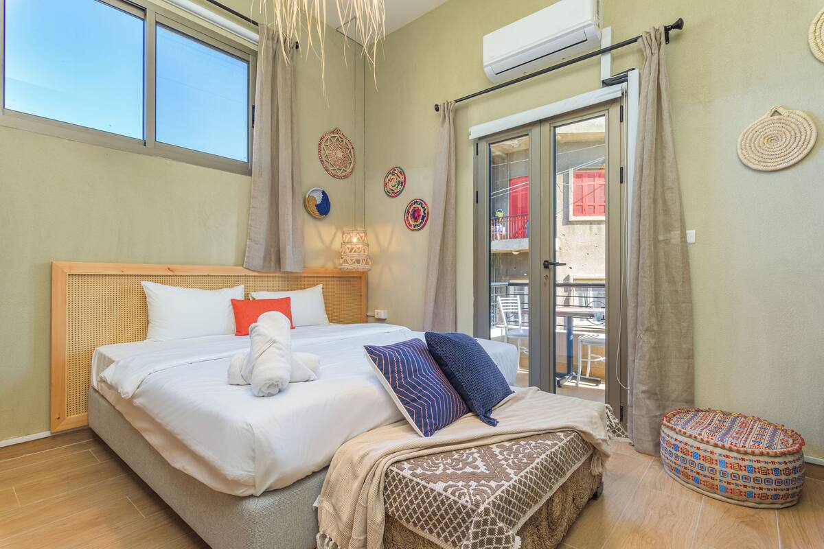A hotel room with a large bed, a small round ottoman, a balcony with a view, and decorative wall art.