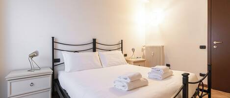 Una cama bien hecha con sábanas y toallas blancas, una mesita de noche con lámpara, un aparador y un radiador.