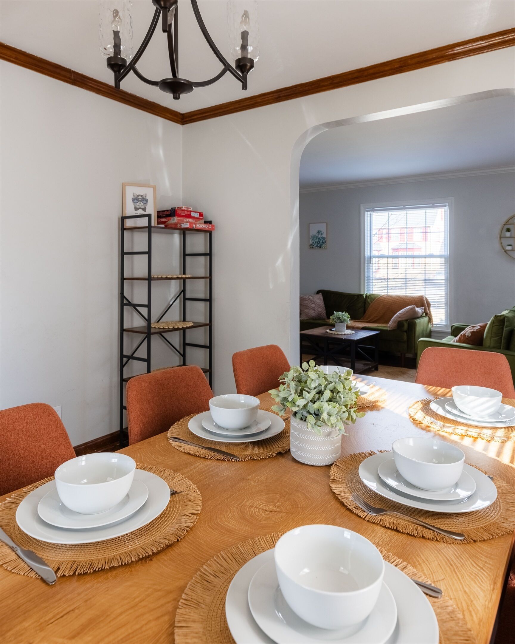 Een eetgedeelte met een houten tafel voor vier personen, omringd door oranje stoelen. Een zwart metalen rek met een fotolijst en een rode doos is links zichtbaar.