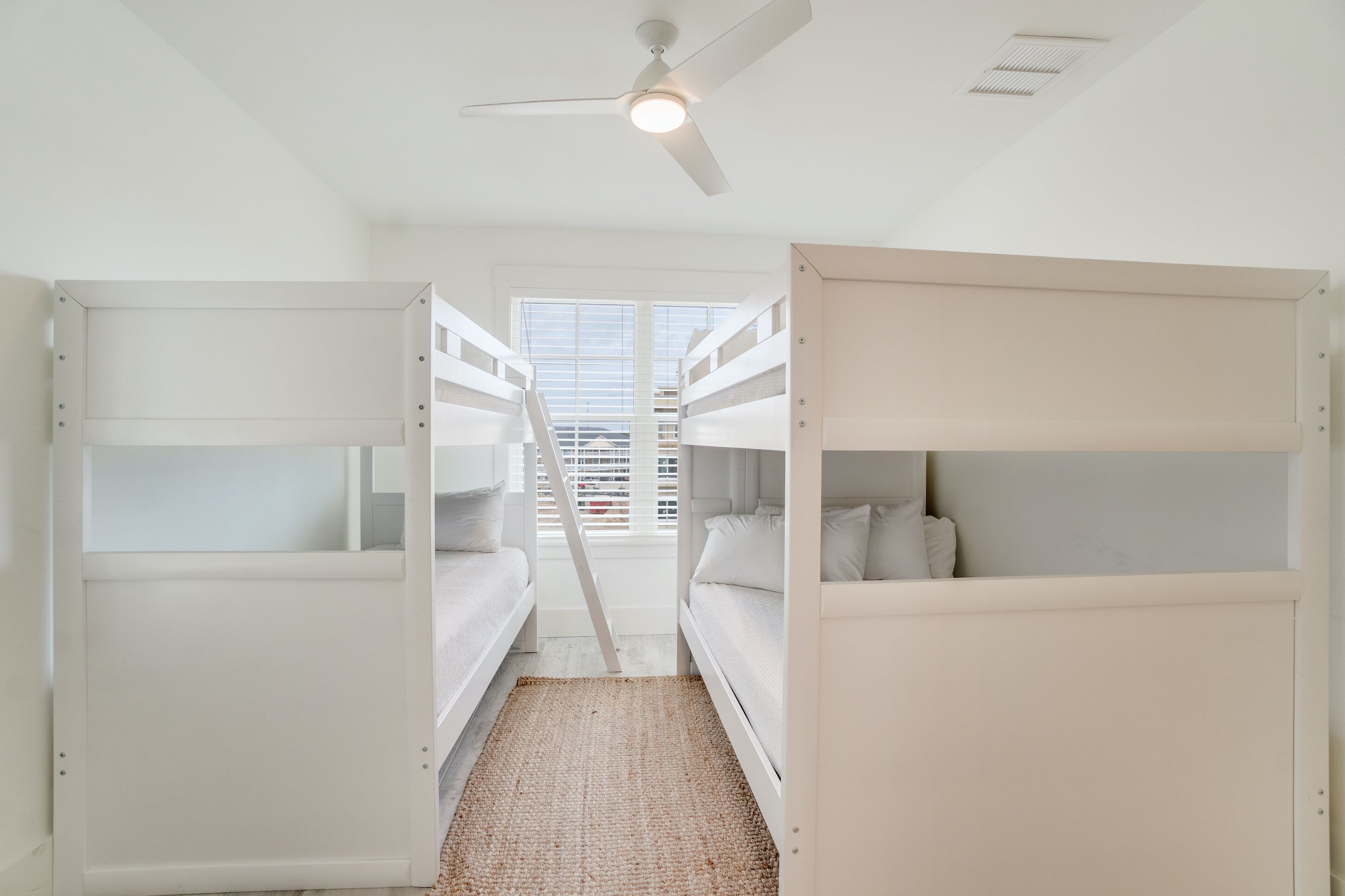 A bunk bed room with a ceiling fan, a window with blinds, and a woven rug on the floor.