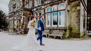 A couple walking in front of a historic stone building with large windows and a bench.