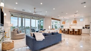A modern living room with a sofa, a coffee table, and a dining area with chairs and a kitchen in the background. - New House, Waterfront, Pool, Hot Tub, Golf Cart (Indian Rocks Beach)