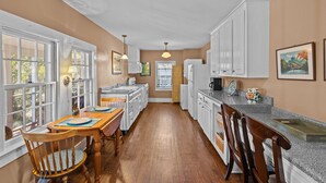 A kitchen with white cabinets, a wooden dining table with chairs, and a window with a view of trees. - Historic District: Downtown New Bern (New Bern)
