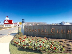 A Coastal Club sign with a speed limit sign of 25 mph, a lighthouse, and a flower bed in front.