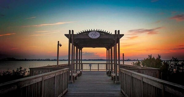 A wooden pier leading to a gazebo with a circular emblem, overlooking a body of water at sunset.