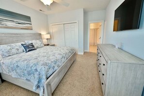 A bedroom with a bed, a dresser, a nightstand, a lamp, a painting, and a door leading to another room.