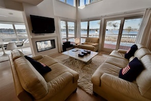 A living room with a fireplace, a television, and a large window overlooking a body of water. - Bethany Beach Waterfront House w Stunning Views * (Bethany Beach)