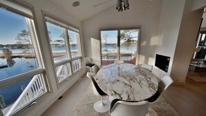 A dining area with a marble table, white chairs, large windows with a view of a lake, and a TV on the wall. - Bethany Beach Waterfront House w Stunning Views * (Bethany Beach)