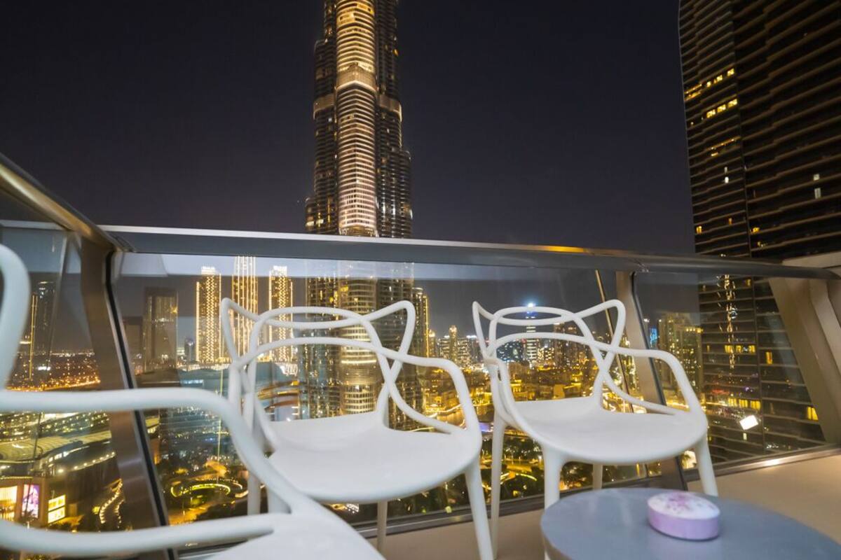A balcony with two white chairs and a small table, overlooking a cityscape with illuminated buildings at night.