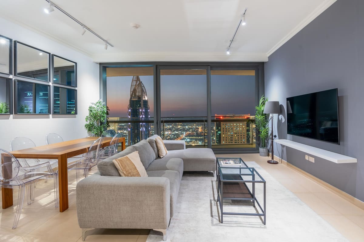 A modern living room with a large window, a grey sofa, a dining table with chairs, and a flat-screen TV mounted on the wall.