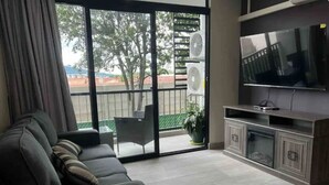 Ein Wohnzimmer mit einer Couch, einem Flachbildfernseher, einer Schiebetür zu einem Balkon und einer Topfpflanze.