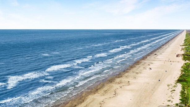 Une longue plage de sable fin, où les vagues viennent se briser sur le rivage, et un ciel bleu et dégagé au-dessus.