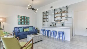 A modern living room with a blue sofa, a coffee table, and a kitchen area with stools and shelves. - Coastal Paradise | 2 Bedrooms | Pool | by Rose (Galveston)