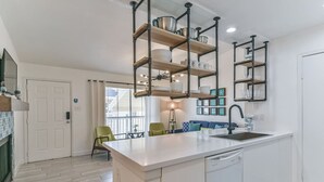 A modern kitchen with white cabinetry, a central island with a sink, and exposed pipe shelves above. - Coastal Paradise | 2 Bedrooms | Pool | by Rose (Galveston)