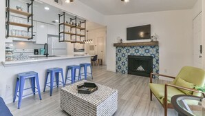 A modern living room with a kitchen area, a fireplace, and a TV mounted above it. - Coastal Paradise | 2 Bedrooms | Pool | by Rose (Galveston)