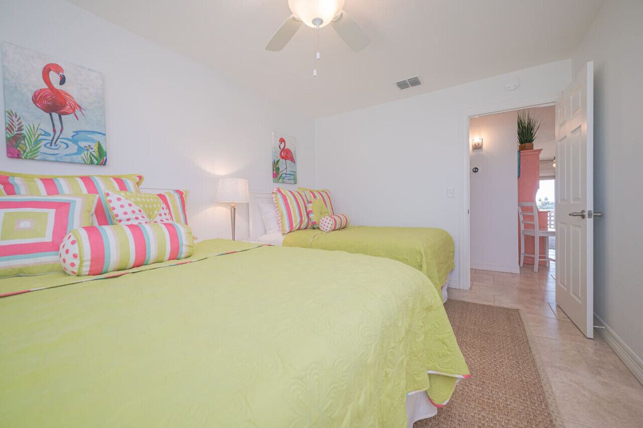 Une chambre avec deux lits, chacun recouvert d’une couverture verte et agrémenté de coussins colorés. On y trouve un ventilateur de plafond, un tableau représentant un flamant rose et une porte donnant sur une autre pièce.