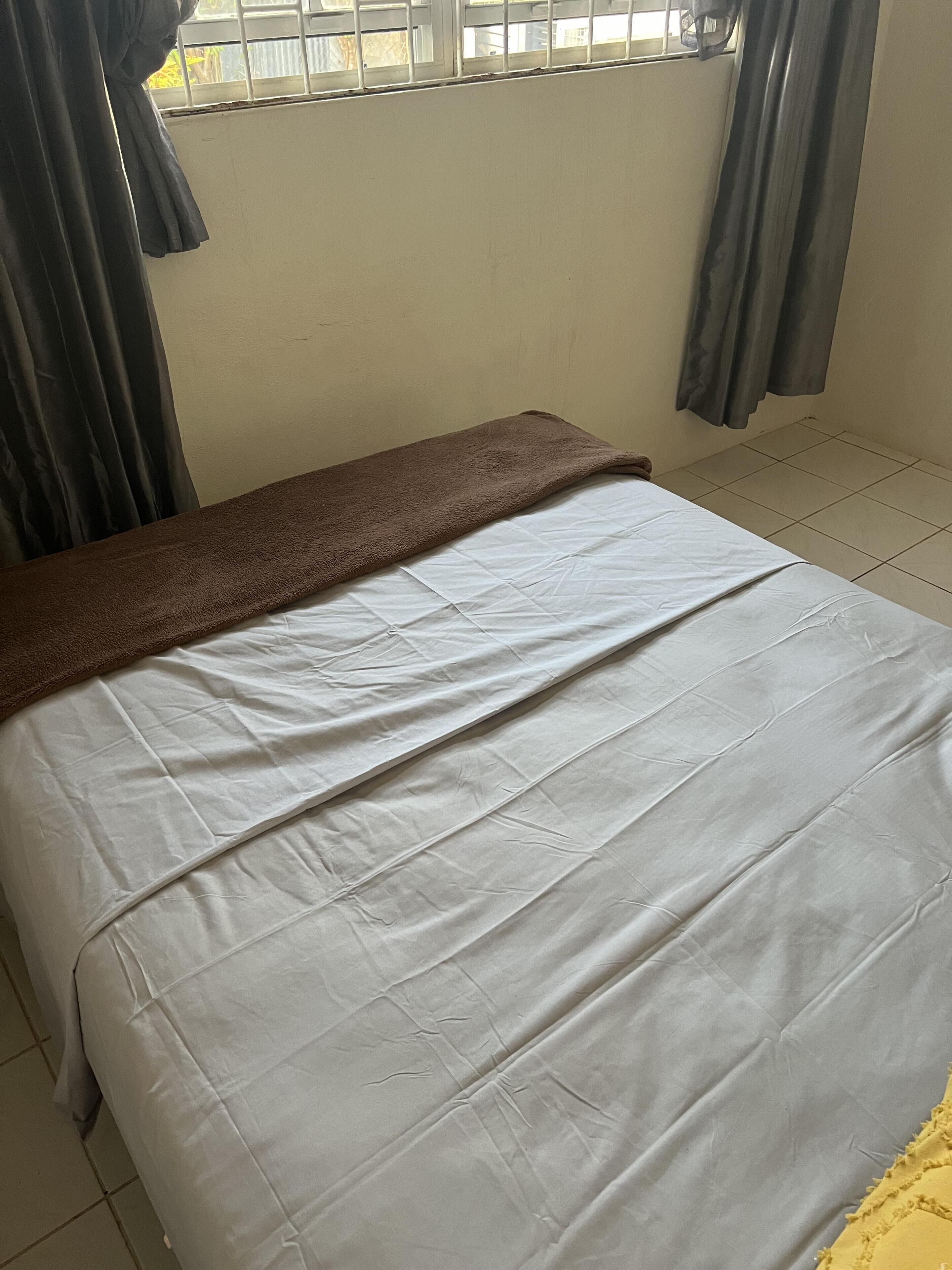A bed with a white sheet and a brown blanket, a window with metal bars, and a tiled floor.