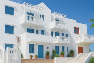 A multi-story white building with blue doors and windows, a balcony with white balustrades, and potted plants at the entrance.