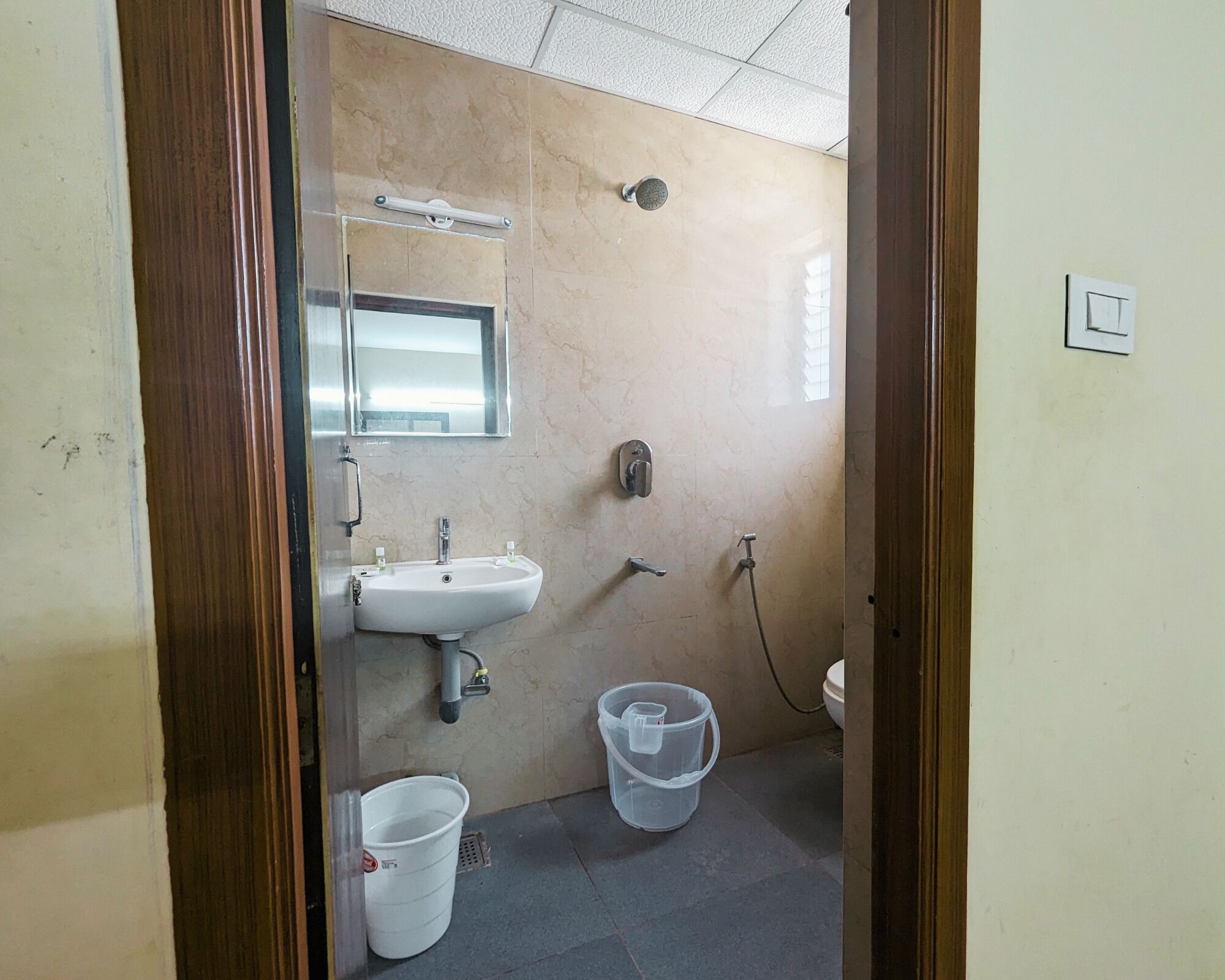 A small bathroom with a sink, mirror, and shower. There are two buckets and a toilet visible.
