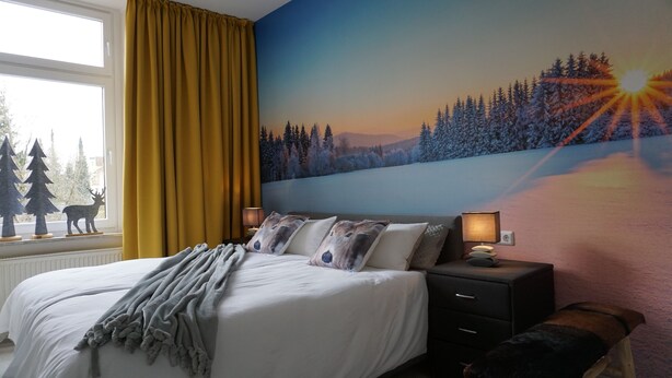 A bedroom with a bed, a nightstand, a bench, a window with curtains, and a wall mural of a snowy landscape.